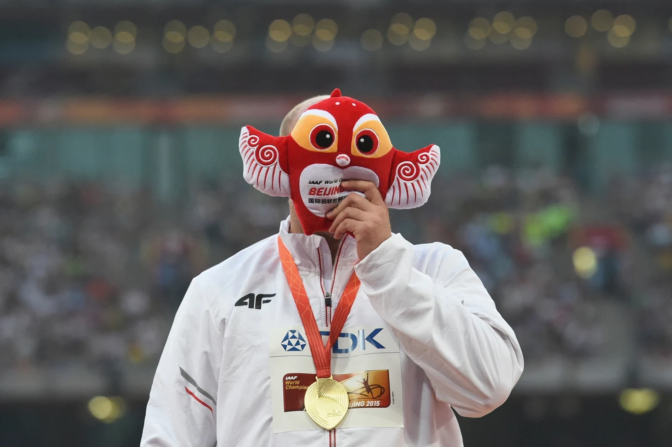 Piotra Małachowski po złotym medalu mistrzostw świata w Pekinie Sportowiec w białym dresie trzyma maskę na twarzy oraz złoty medal na szyi, na tle stadionu wypełnionego widownią