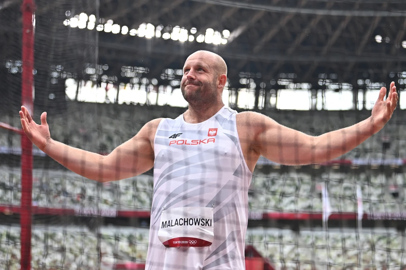 Atleta w stroju reprezentacji Polski podczas zawodów lekkoatletycznych unosi ręce w geście triumfu lub dumy, w tle stadion wypełniony pustymi trybunami.