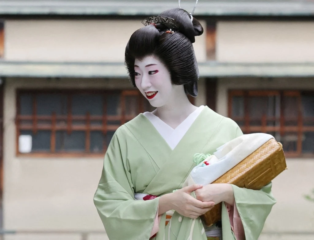 Kobieta w tradycyjnym japońskim stroju kimono o jasnej barwie, z misternie ułożonymi włosami, starannym makijażem i delikatnym uśmiechem, trzymająca w rękach ozdobny przedmiot lub torebkę, na tle budynku o drewnianych oknach.