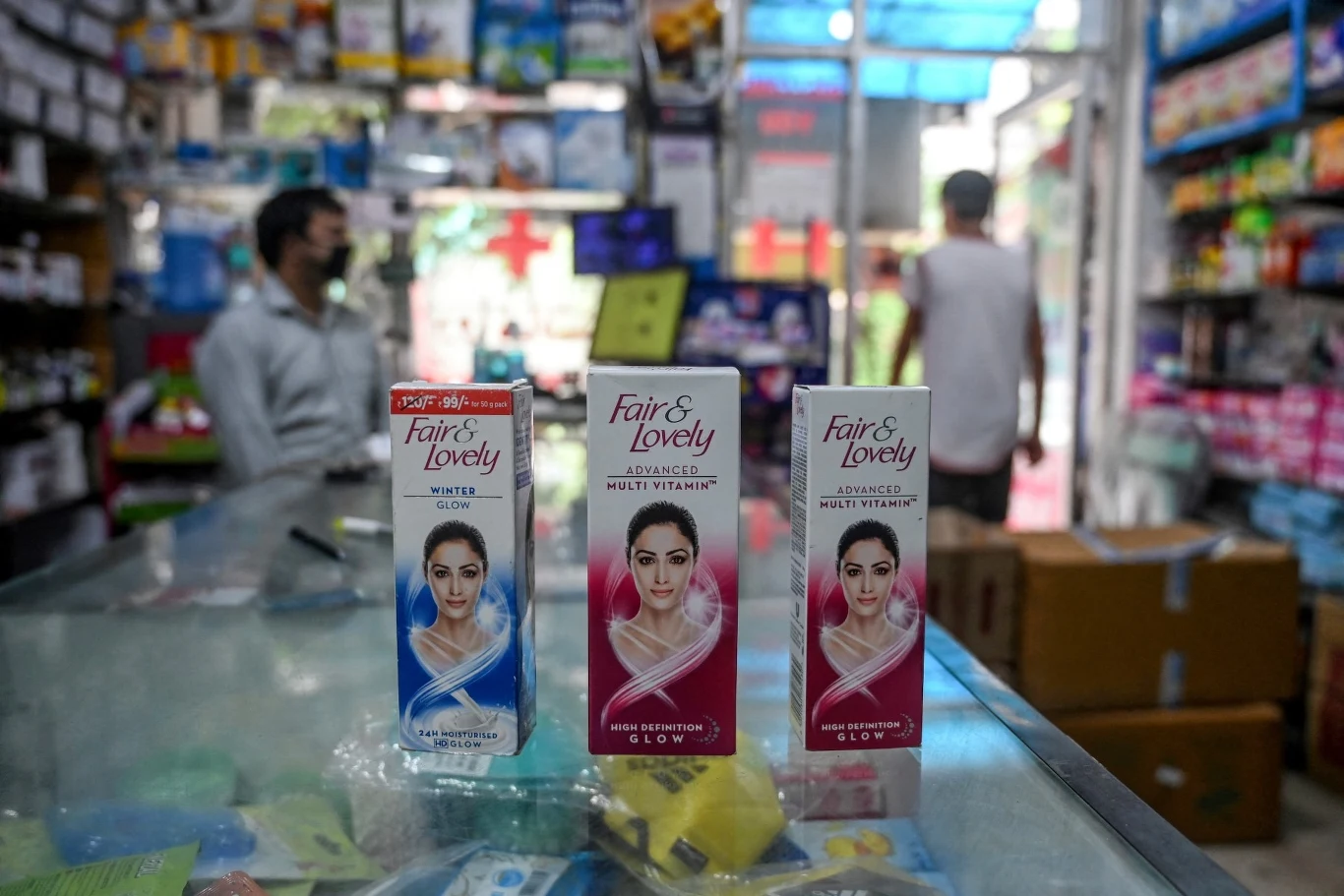 Trzy kremy marki Fair & Lovely ustawione na szklanej ladzie w aptece lub sklepie, w tle widoczne półki z różnymi produktami oraz dwie osoby, z których jedna ma założoną maseczkę ochronną.