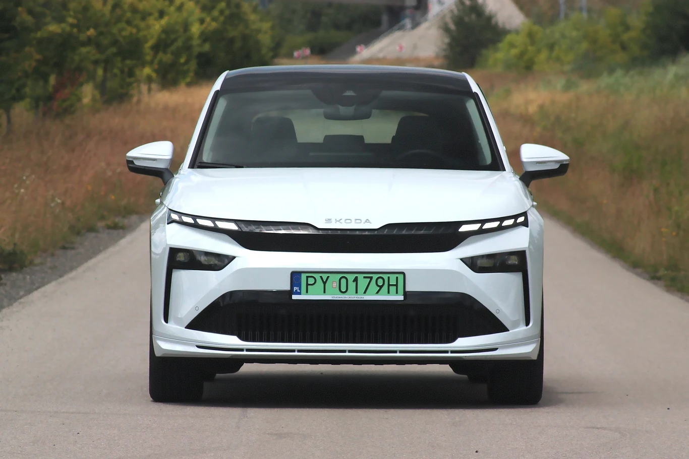 Biały samochód marki Skoda, widziany z przodu, stoi na asfaltowej drodze otoczonej zielenią, z widoczną tablicą rejestracyjną PY 0179H.