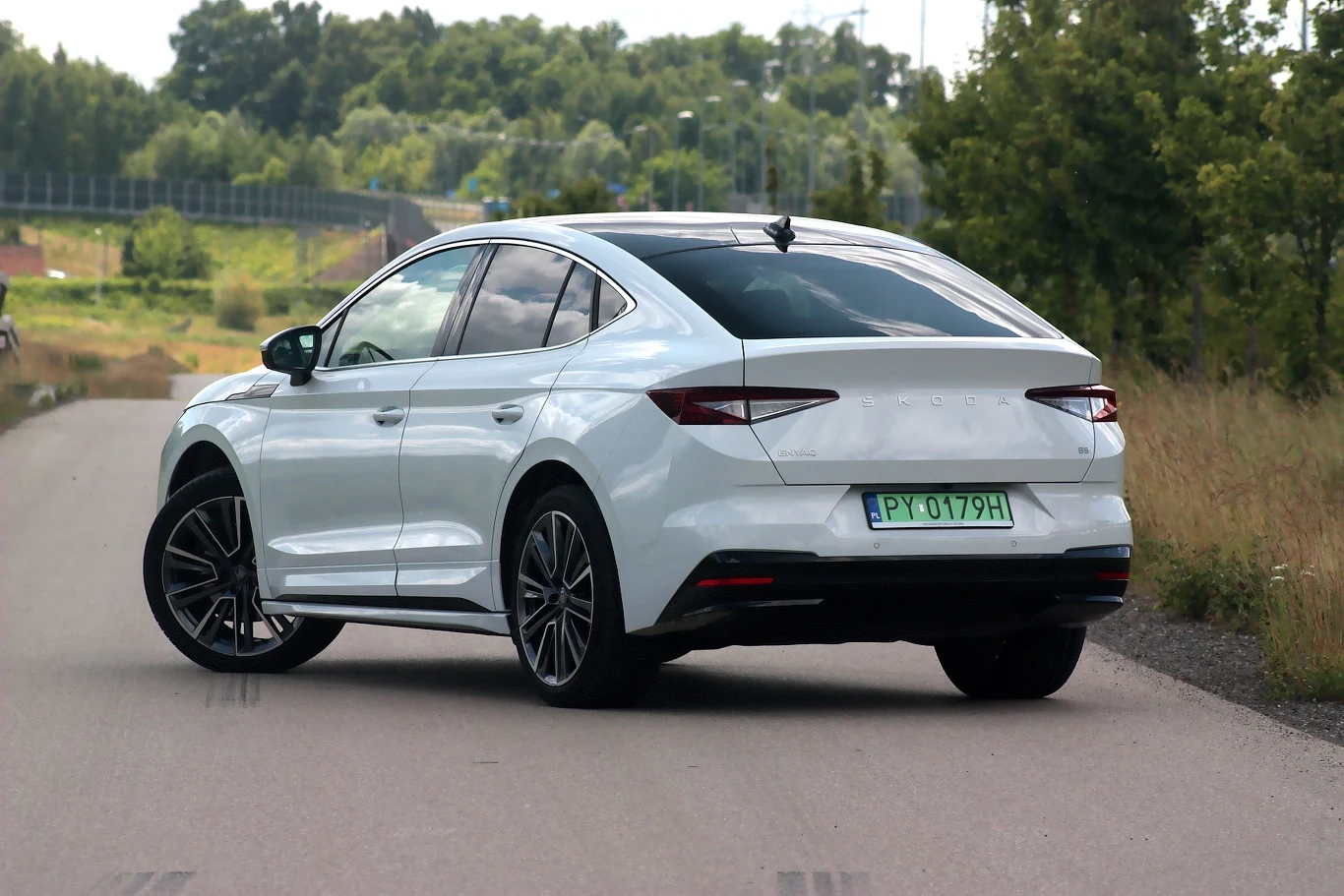 Białe auto typu liftback jedzie pustą, asfaltową drogą otoczoną zielenią, widoczne są nowoczesne detale nadwozia oraz tablica rejestracyjna.