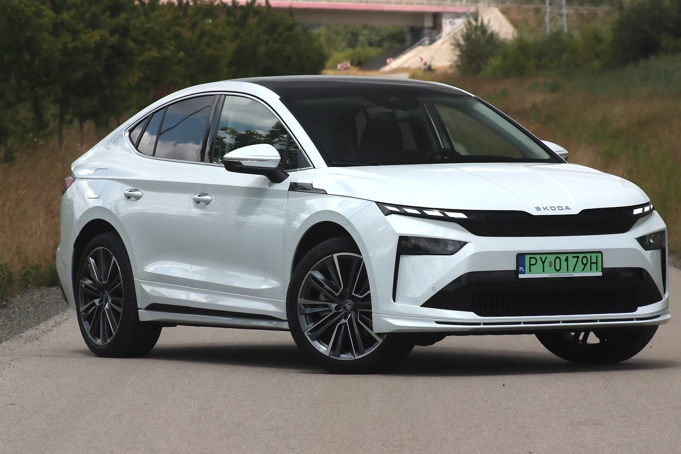 Nowoczesny, biały samochód marki Škoda z zieloną tablicą rejestracyjną stoi na bocznej, asfaltowej drodze. Otoczenie stanowi zieleń oraz niewielki most w tle.