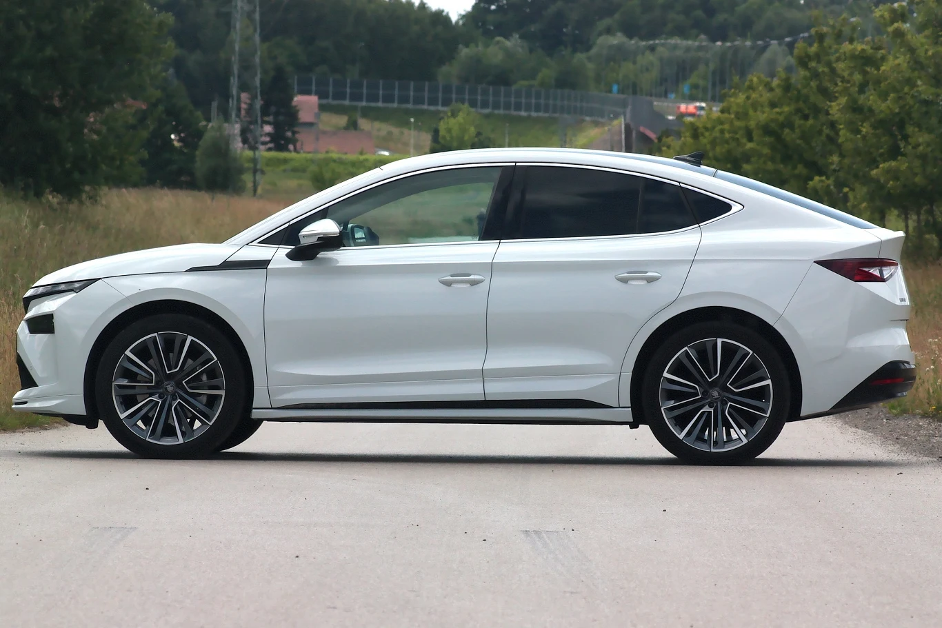 Biały samochód osobowy klasy średniej lub wyższej zaparkowany przy drodze, nowoczesna sylwetka z aerodynamicznymi liniami i dużymi alufelgami, otoczenie stanowi zielona roślinność oraz droga asfaltowa