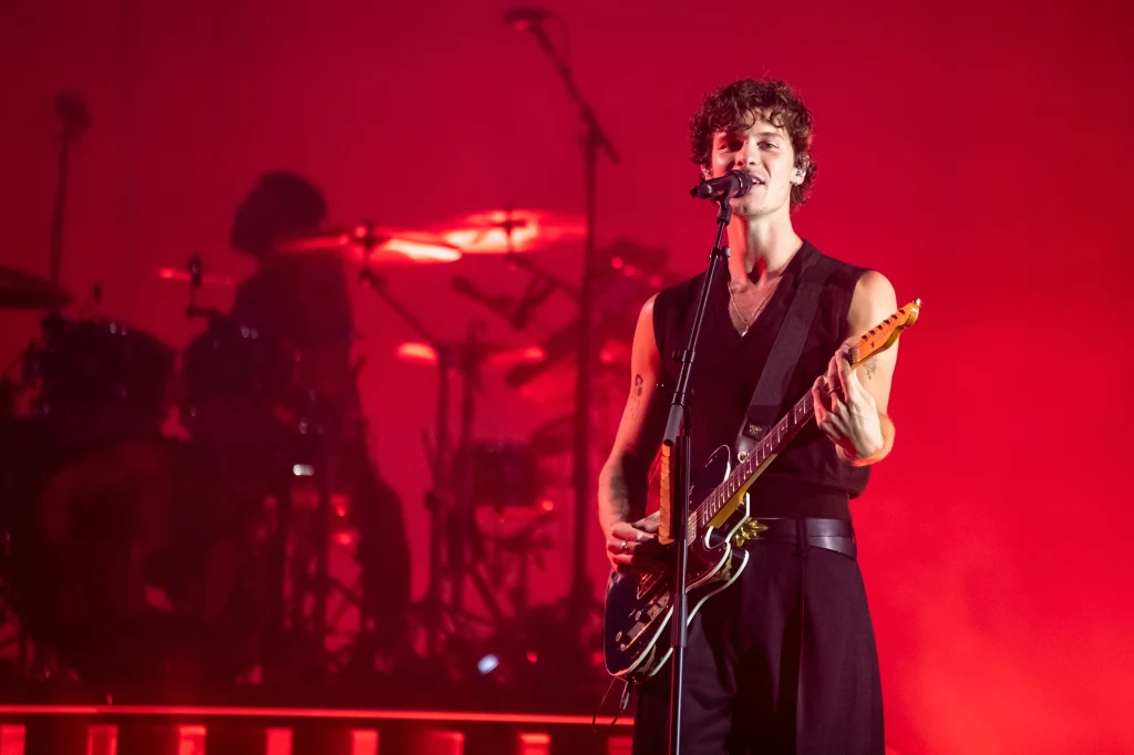 Wokalista grający na gitarze elektrycznej podczas koncertu, oświetlony intensywnym czerwonym światłem, w tle widoczny rozmyty perkusista i elementy sceny.