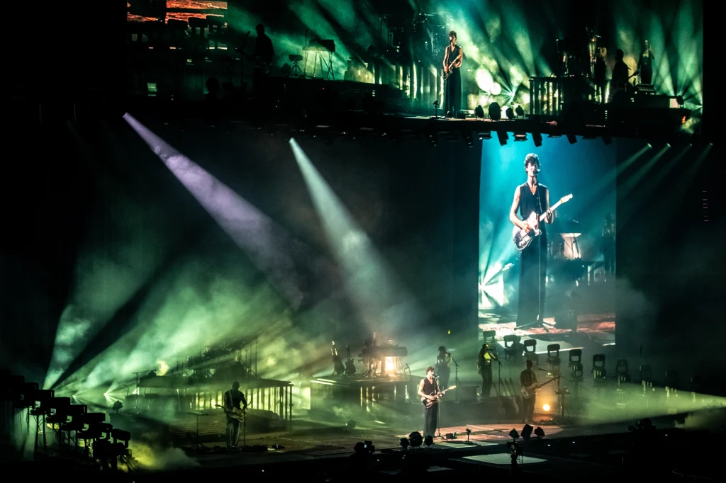 Koncert rockowy z efektownym oświetleniem, w centrum sceny znajduje się gitarzysta, którego sylwetka jest wyraźnie widoczna zarówno na scenie, jak i na dużym ekranie z boku, wokół rozstawiona jest reszta zespołu i sprzęt muzyczny, a całość podkreślają ...