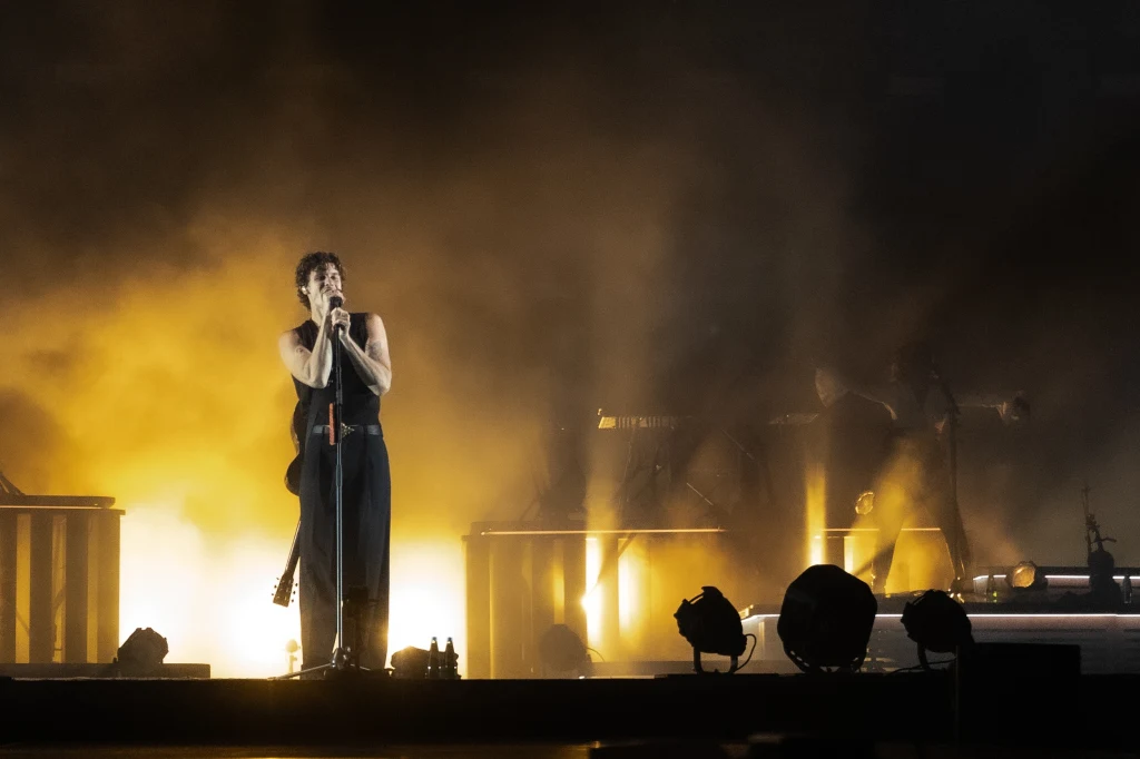 Wokalista na scenie oświetlonej dynamicznymi światłami, stojący przy mikrofonie z gitarą na plecach, w tle widoczna scenografia oraz sprzęt muzyczny, atmosfera koncertowa podkreślona przez intensywne dymy i światła.