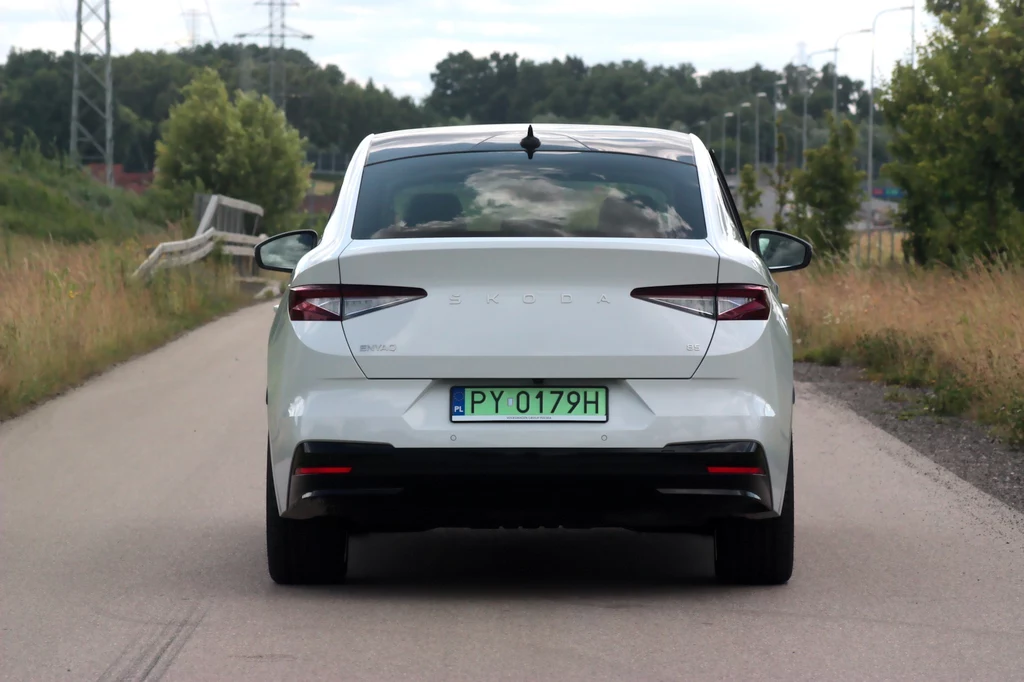 Skoda Enyaq Coupe z tyłu nie wygląda zbyt atrakcyjnie Biały samochód osobowy marki Skoda o numerze rejestracyjnym PY 0179H jedzie asfaltową drogą, otoczoną przez zieleń oraz niewysokie drzewa. Widoczna tylna szyba auta z bagażem lub przewożonymi przedmiotami, brak innych pojazdów na drodze.