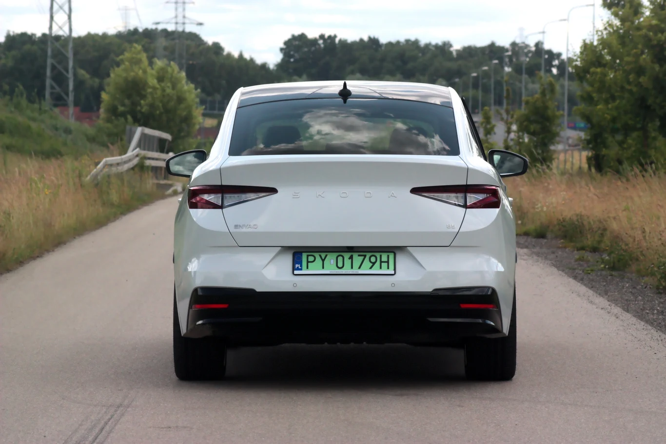 Biały samochód osobowy marki Skoda o numerze rejestracyjnym PY 0179H jedzie asfaltową drogą, otoczoną przez zieleń oraz niewysokie drzewa. Widoczna tylna szyba auta z bagażem lub przewożonymi przedmiotami, brak innych pojazdów na drodze.