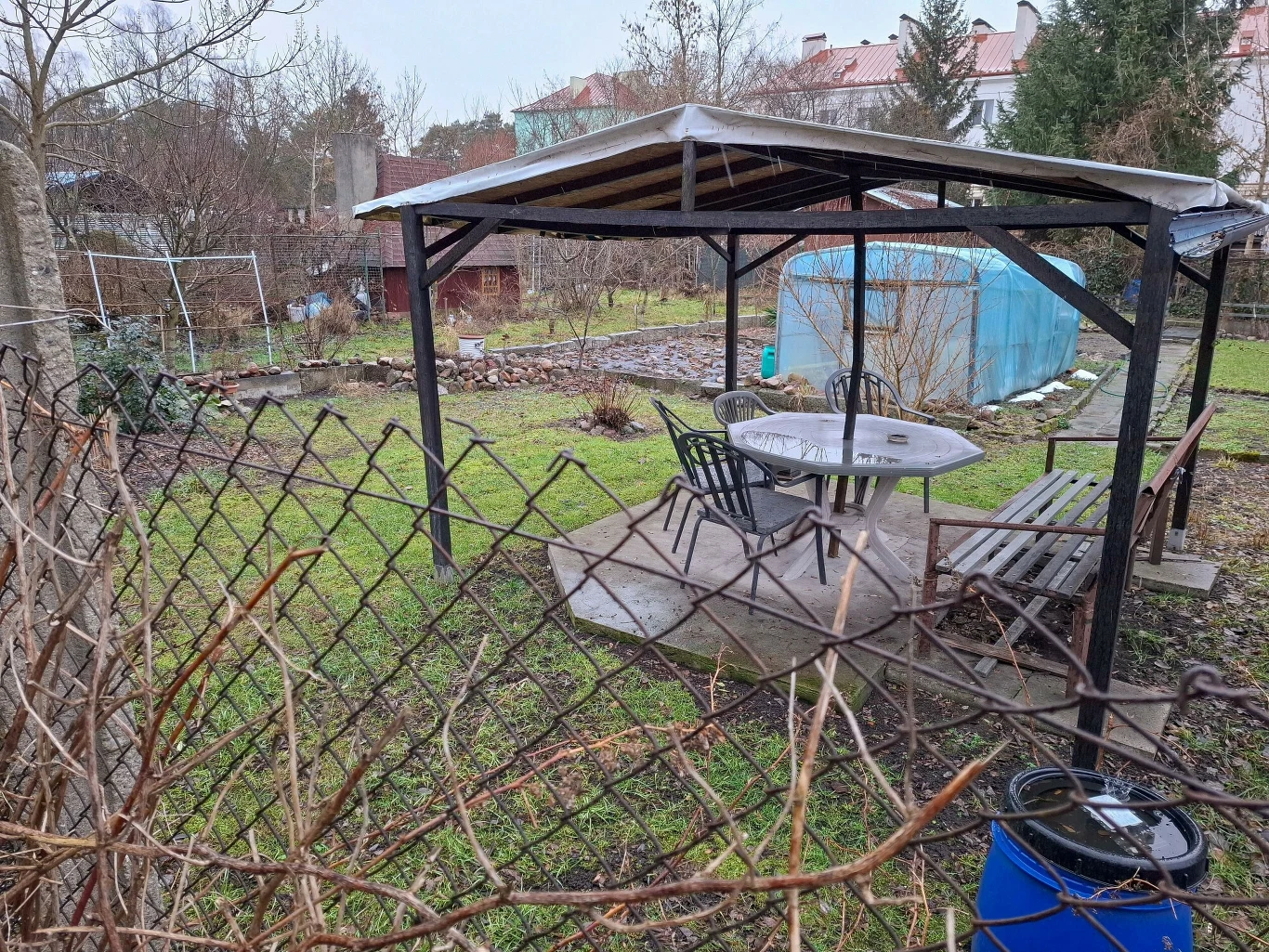 Altana ogrodowa z metalowym dachem, plastikowy stół i cztery krzesła, obok ławka. W tle szklarnia oraz ogrodzone działki z roślinnością i sąsiadującymi domami.