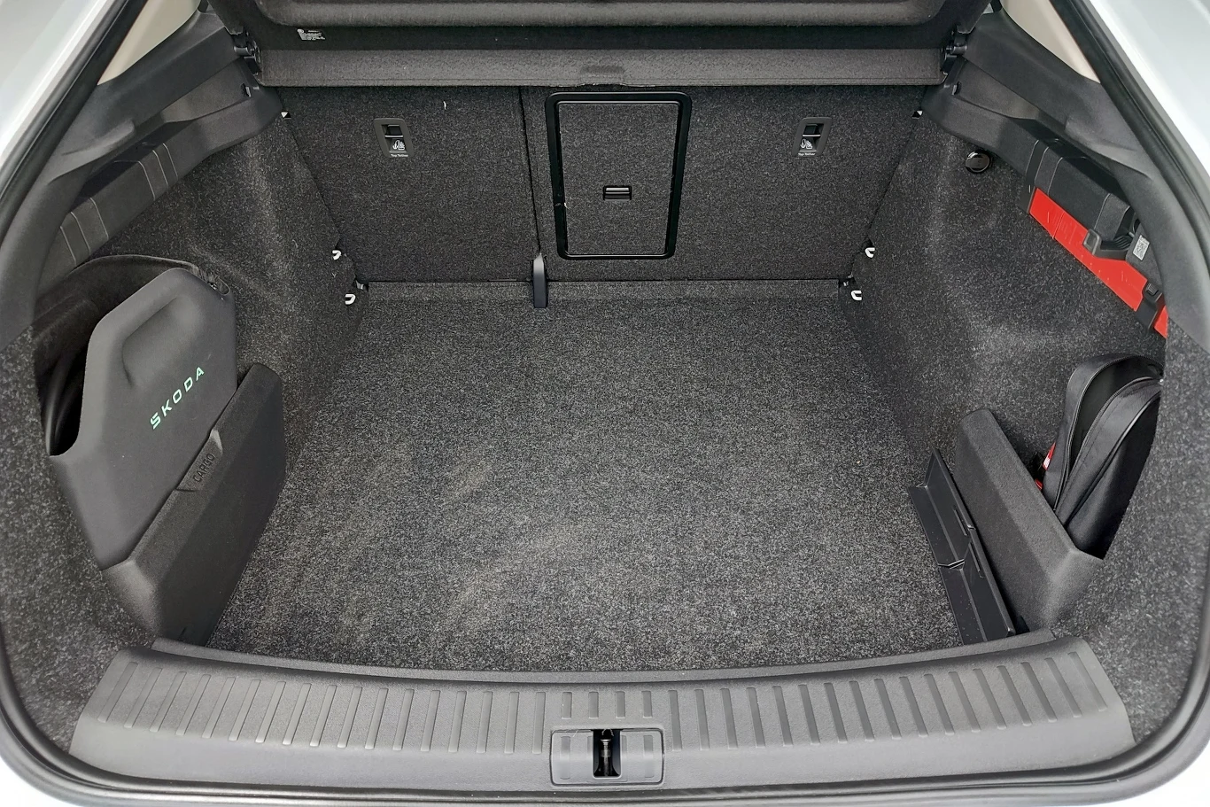 Pojemny bagażnik samochodu wyłożony szarą wykładziną, po bokach schowki na akcesoria i widoczne mocowania, w tle otwór na narty.