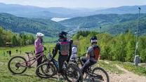 Grupa rowerzystów w kaskach i ochraniaczach zatrzymuje się na szutrowej ścieżce na zboczu góry, za plecami rozległy górski krajobraz z jeziorem w oddali, wokół zielone lasy i łagodne wzgórza.