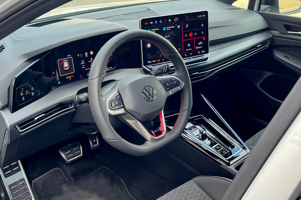 Sportowa kierownica z perforowaną skórą, dekory imitujące włókno węglowe oraz nowy system multimedialny. Nowoczesne wnętrze samochodu marki Volkswagen z wyraźnie widocznymi dwoma dużymi ekranami wielofunkcyjnymi, sportową kierownicą z logotypem VW oraz estetyczną konsolą środkową ze skrzynią biegów o designerskim kształcie i chromowanymi wykończeniami.