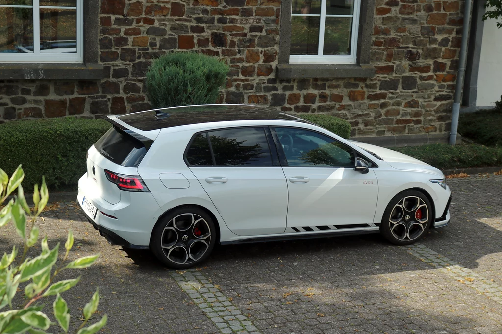 Wersja Clubsport wyróżnia m.in. sporej wielkości spojlerem nad tylną szybą. Biały samochód osobowy Volkswagen Golf GTI z czarnym dachem i sportowymi felgami zaparkowany przy brukowanej alejce, w pobliżu budynku z ceglaną elewacją i ozdobnymi krzewami.