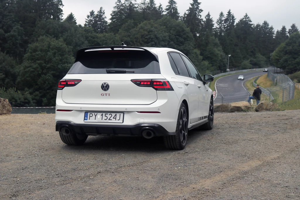 Tor Nürburgring Nordschleife nie bez powodu nazywany jest "Zielonym Piekłem" Biały Volkswagen Golf GTI z polską rejestracją stoi tyłem na żwirowym placu, w tle widać zakręt toru wyścigowego i zalesioną okolicę.