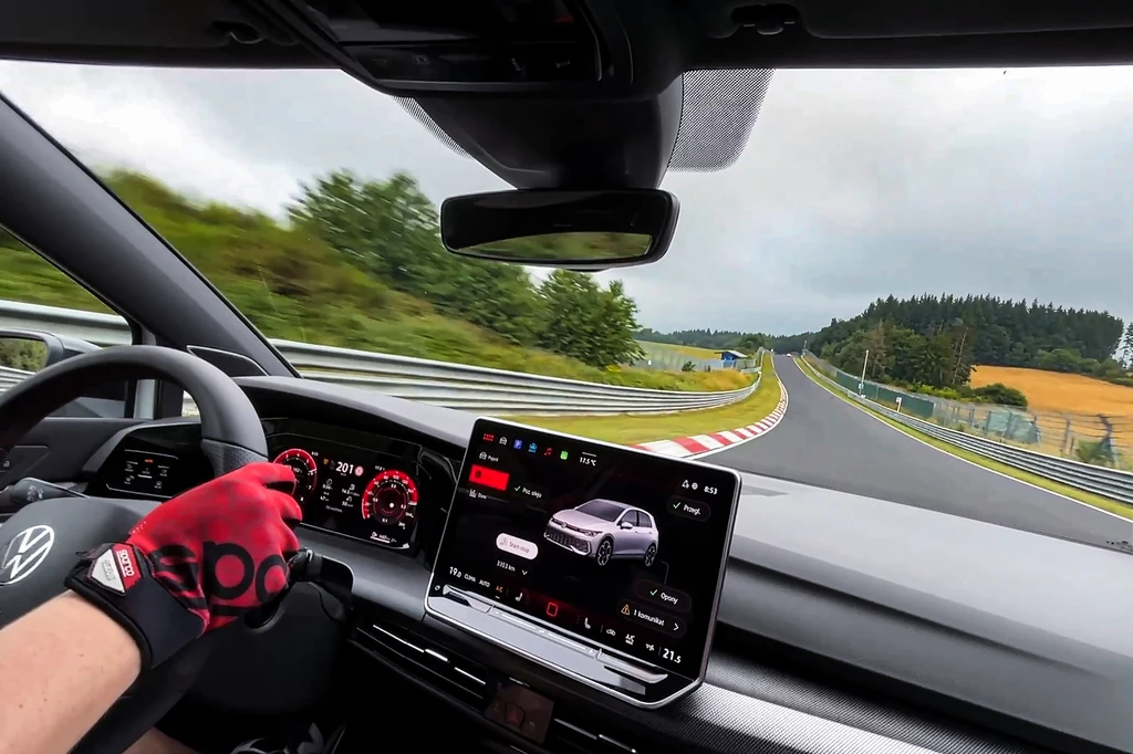 W rejonie łuku Schwedenkreuz, prędkości są już naprawdę wysokie. Kierowca w sportowych rękawiczkach prowadzi samochód marki Volkswagen po torze wyścigowym, wnętrze auta z widocznym cyfrowym kokpitem i dużym ekranem multimedialnym, za szybą dynamiczny zakręt otoczony zielenią i barierkami.