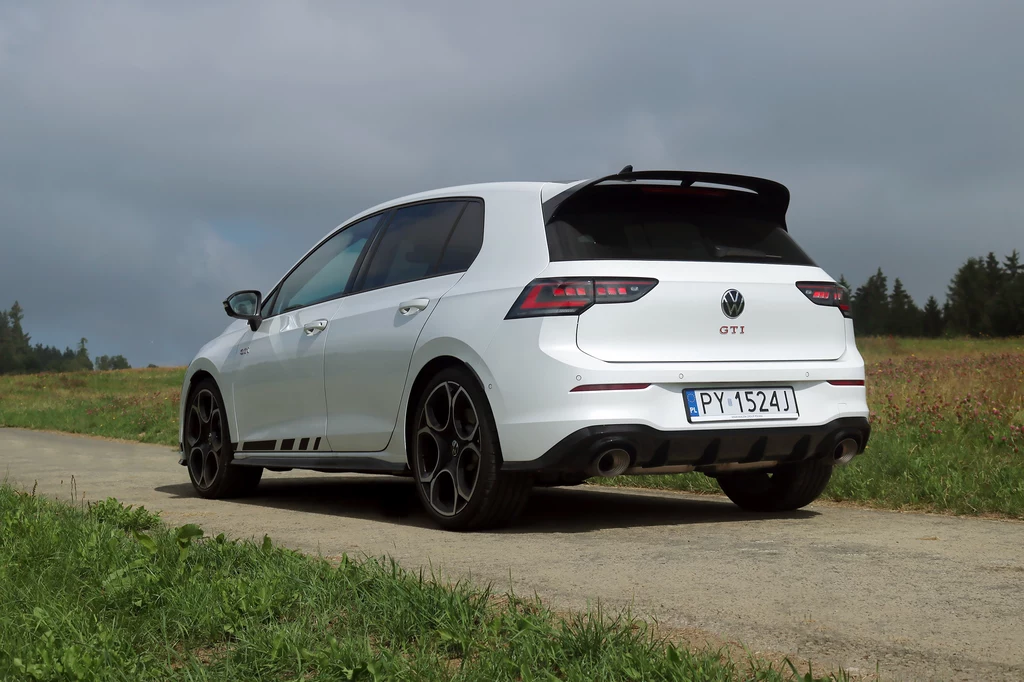 Golf GTI Clubsport przeszedł istotne zmiany. Zawieszenie zostało utwardzone i obniżone o 10 mm. Biały sportowy samochód Volkswagen Golf GTI zaparkowany na wąskiej asfaltowej drodze, wokół rozciągają się zielone łąki i drzewa, na niebie widoczne zachmurzenie.