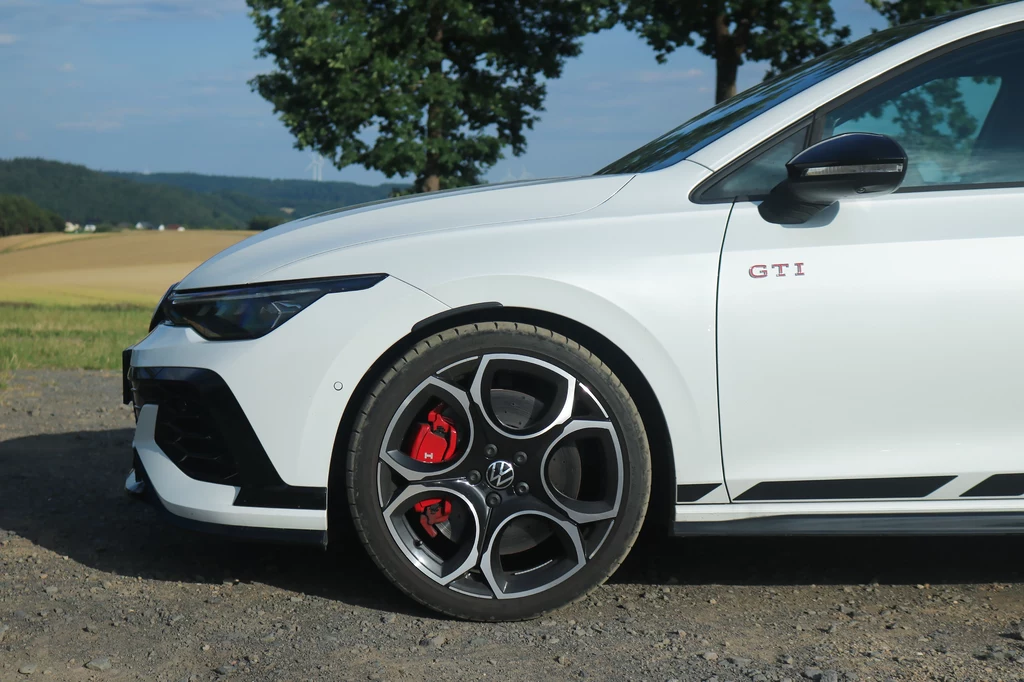 Po całym okrążeniu układ hamulcowy nadal działa bez zarzutu. Przednia część białego samochodu sportowego Volkswagen GTI z charakterystycznym napisem GTI na nadkolu, nowoczesnymi aluminiowymi felgami i czerwonymi zaciskami hamulców, na tle wiejskiego krajobrazu.