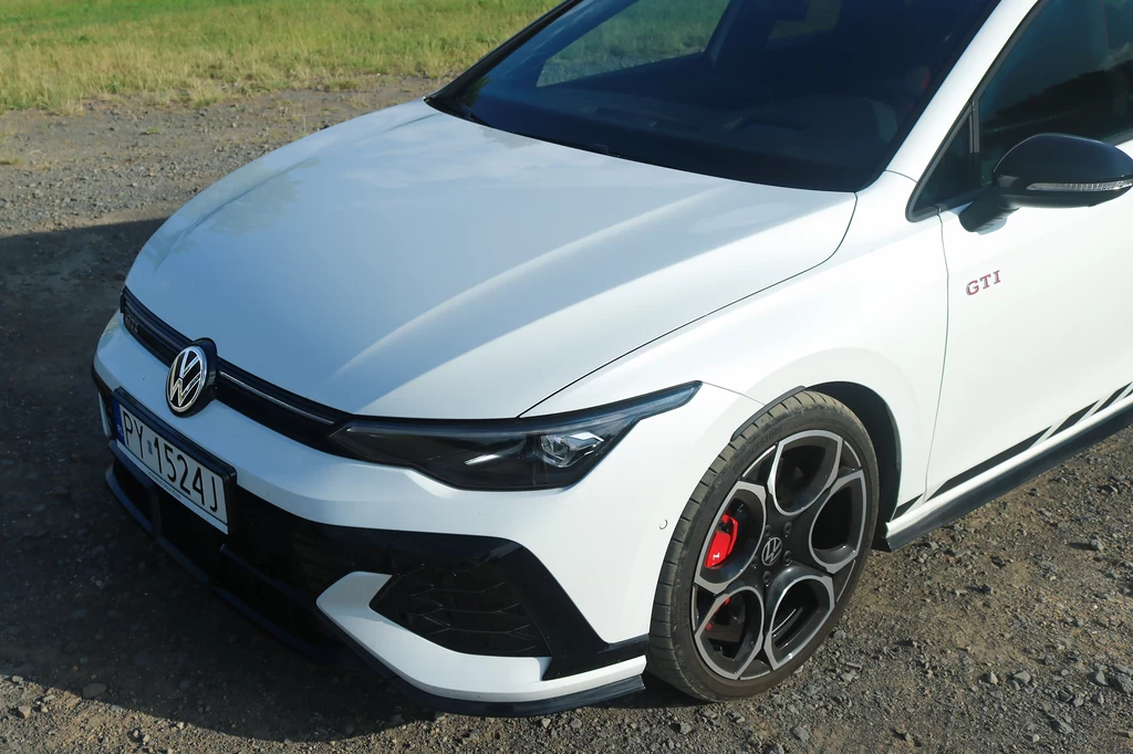 Bystre oko dostrzeże także subtelne zmiany w przednim zderzaku. Przód białego samochodu Volkswagen Golf GTI z charakterystycznym logo, sportowymi felgami i czerwonymi akcentami, zaparkowany na żwirowym podłożu w słoneczny dzień.