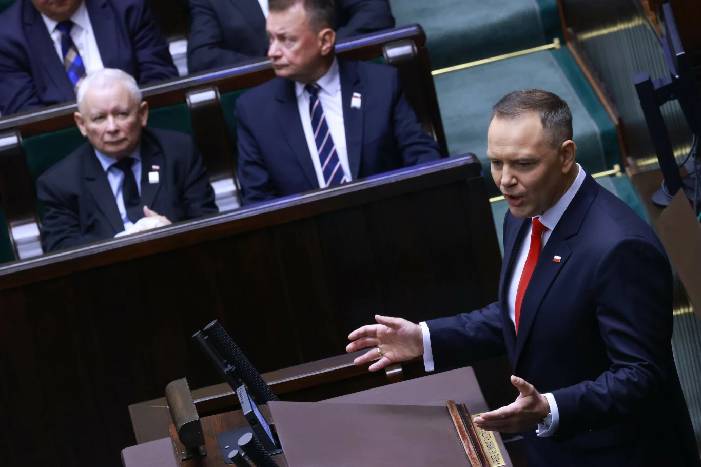 Prezydent Karol Nawrocki podczas przemówienia w Sejmie po zaprzysiężeniu Prezydent przemawia z mównicy sejmowej, ujęty podczas gestykulacji; w tle siedzą politycy w garniturach, obserwujący wystąpienie.