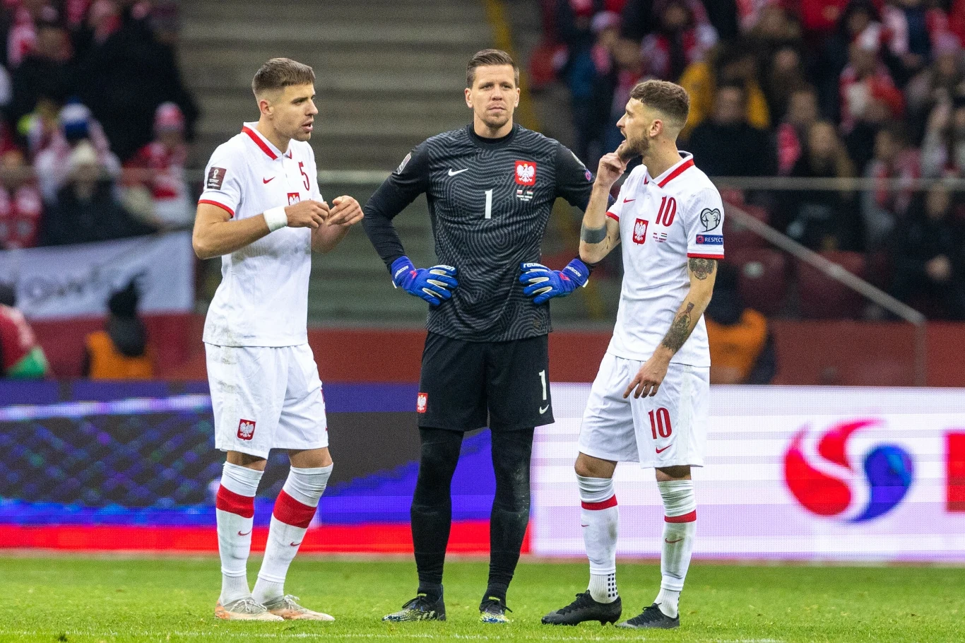 Trzech polskich piłkarzy w strojach reprezentacyjnych rozmawia na murawie stadionu podczas meczu, z widoczną publicznością na trybunach w tle.