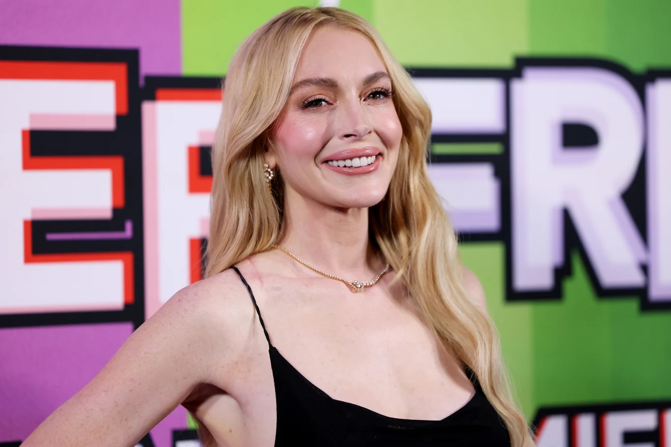 Lindsay Lohan zachwyciła fanów swoim wyglądem Uśmiechnięta kobieta o długich blond włosach w czarnej sukience na tle kolorowej ściany z graficznymi napisami.