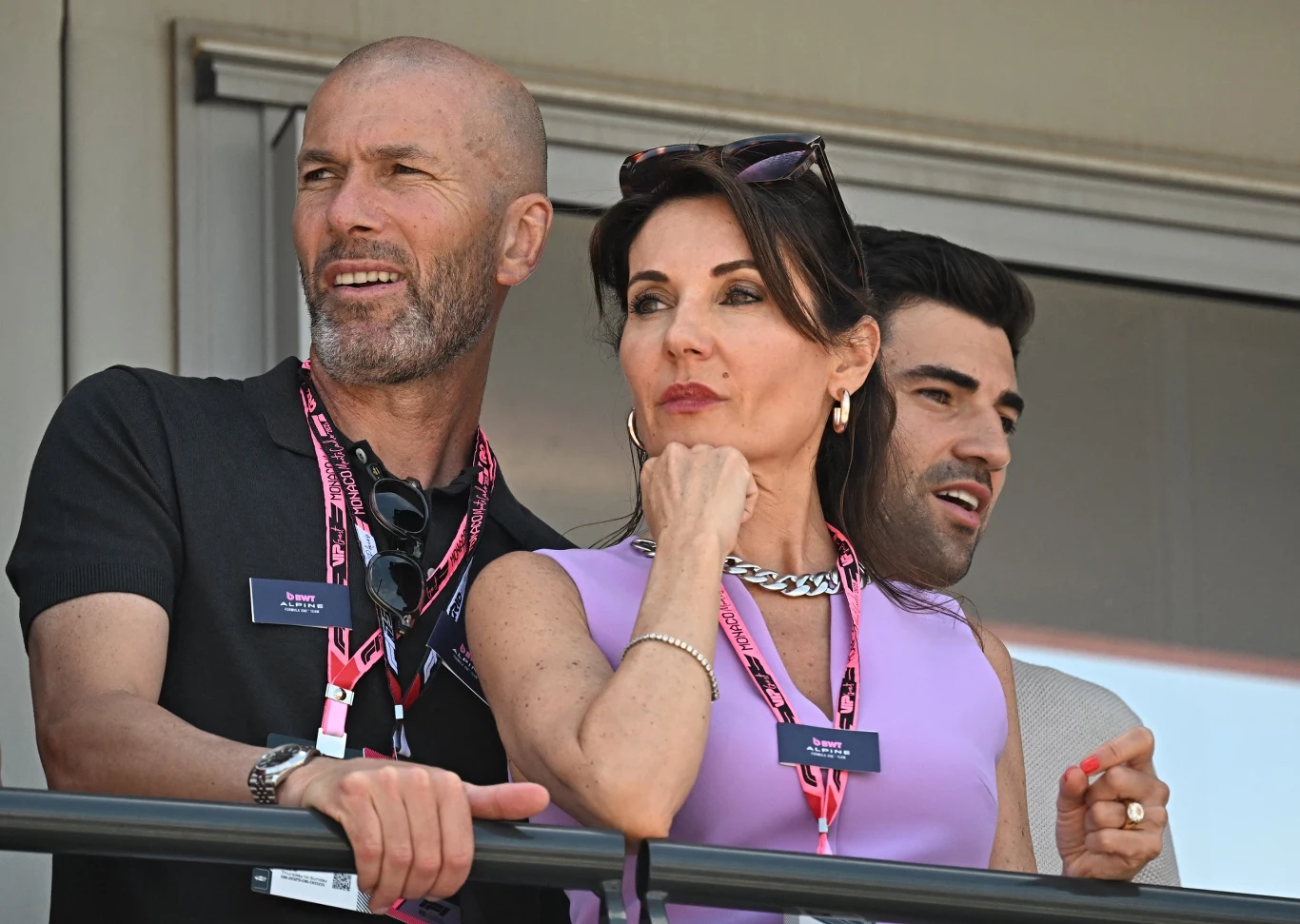 Zinedine Zidane w towarzystwie małżonki Troje dorosłych obserwuje coś z balkonu lub tarasu, ubrani elegancko, mają identyfikatory na szyjach, panuje atmosfera podziwu i skupienia.