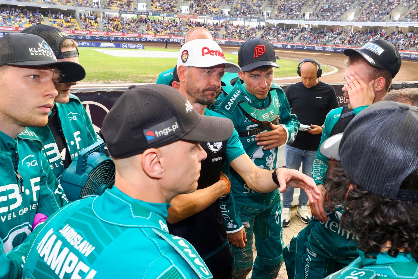 Grupa zawodników żużlowych w zielonych strojach klubowych zgromadzona w kręgu podczas intensywnej narady na stadionie sportowym, wokół liczni kibice na trybunach.