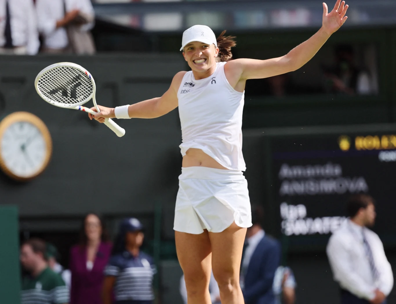 Iga Świątek Tenisistka w białym stroju sportowym skacze z radości na korcie, trzymając rakietę tenisową, w tle widoczne są tablice wyników oraz sędziowie i publiczność.