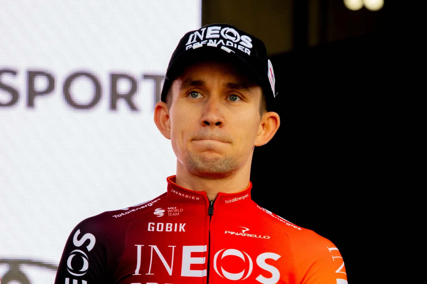 Michał Kwiatkowski Zawodnik kolarski w czerwono-czarnym stroju drużyny Ineos Grenadiers oraz czapce z logo zespołu na tle rozświetlonego ekranu.