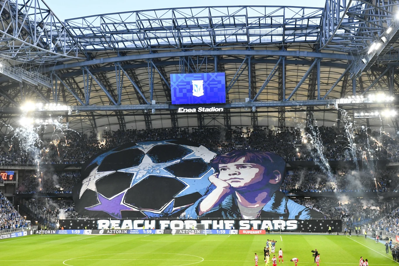 Kibice Lecha Poznań liczą dziś na niespodziankę w meczu z FSV Mainz Tłum kibiców na stadionie piłkarskim tworzy efektowną oprawę z wielkim transparentem, na którym widnieje rysunek dziecka patrzącego w gwiazdy oraz napis 'Reach for the stars', pośrodku widać wielką piłkę stylizowaną na logo Ligi Mistrzów.
