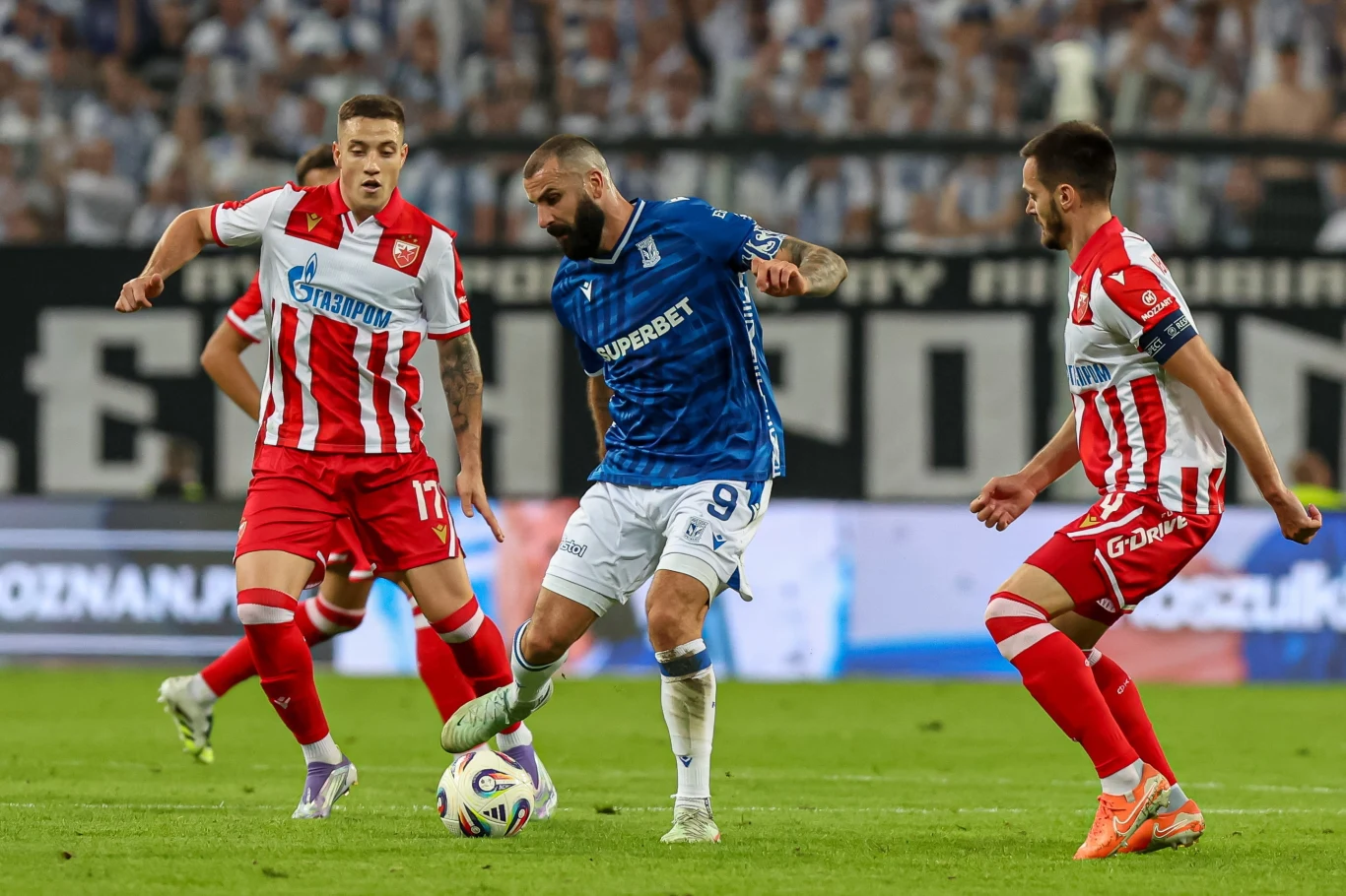 Piłkarz w niebieskiej koszulce prowadzi piłkę nożną, a dwóch zawodników w czerwono-białych strojach próbuje odebrać mu piłkę na tle trybun pełnych kibiców.