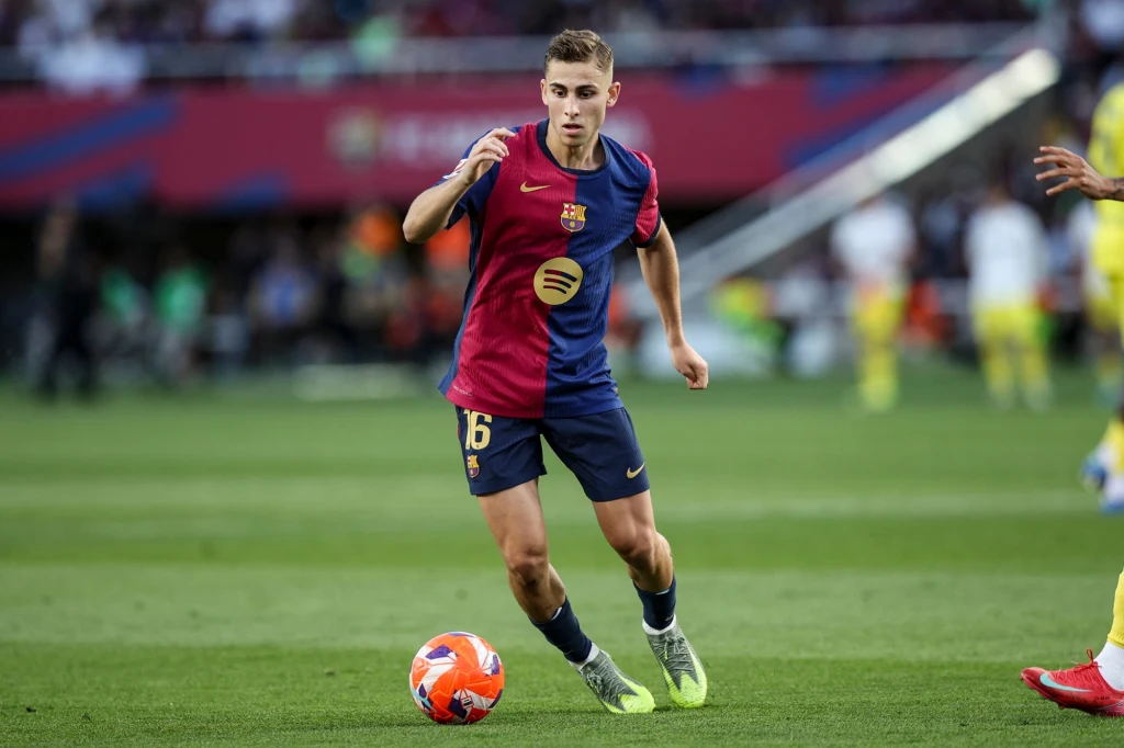 Piłkarz w granatowo-bordowej koszulce FC Barcelony drybluje z piłką na boisku w trakcie meczu, w tle rozmazana publiczność oraz inni zawodnicy.