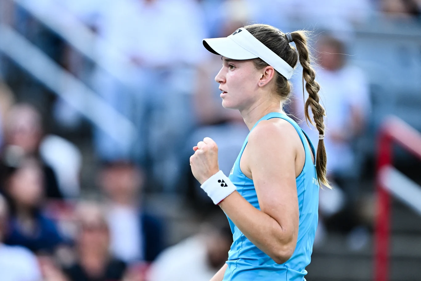 Jelena Rybakina Tenisistka w sportowym stroju podczas meczu na korcie, zebrane włosy w długi warkocz, skupiona wyraz twarzy oraz zaciśnięta pięść sygnalizująca motywację i determinację. W tle rozmazana publiczność.