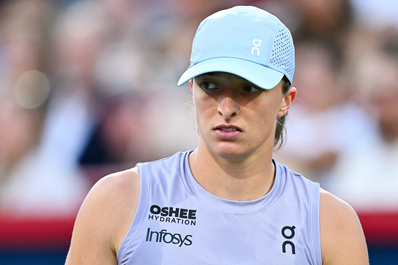 Iga Świątek Młoda kobieta w sportowej czapce i koszulce bez rękawów z widocznymi logotypami sponsorów, skupiona na tle rozmytego stadionu.