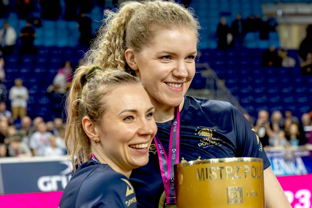 Dwie uśmiechnięte kobiety w sportowych koszulkach świętują zwycięstwo, trzymając złoty puchar z napisem 'Mistrz Polski', w tle widoczne trybuny z kibicami.
