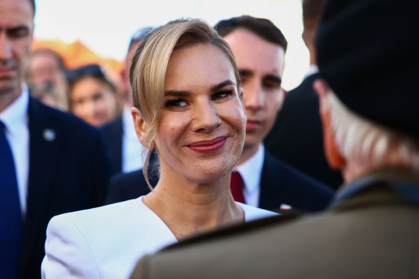 Kobieta w eleganckim białym stroju uśmiecha się, otoczona przez innych ludzi, atmosfera oficjalnego spotkania lub wydarzenia.