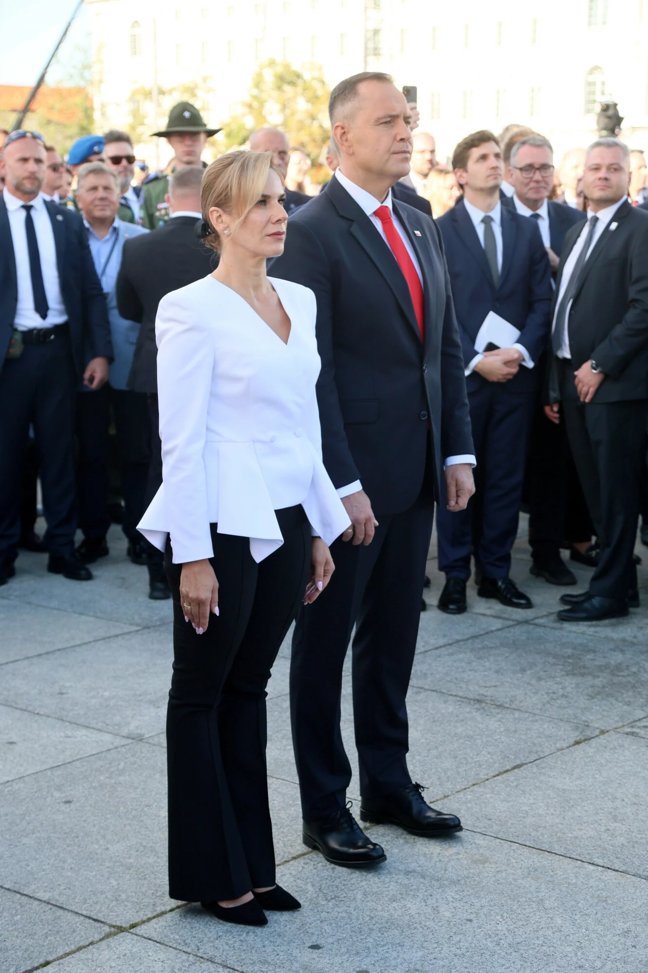 Kobieta w białej marynarce i czarnych spodniach oraz mężczyzna w garniturze z czerwoną krawatą stoją razem na tle grupy elegancko ubranych osób podczas oficjalnej uroczystości na świeżym powietrzu.