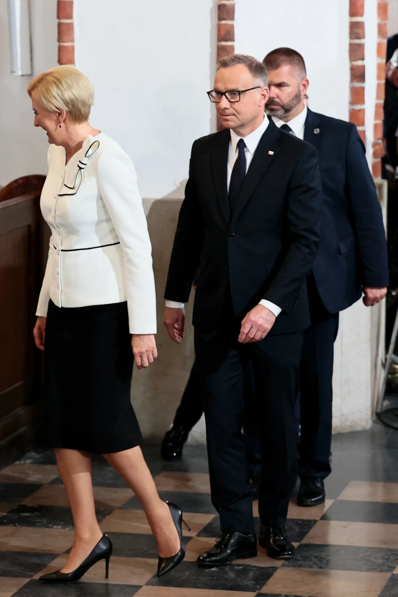 Trzy osoby w formalnych strojach idące marmurową posadzką w eleganckim, historycznym wnętrzu, dwie z nich skupione na przodzie, jedna z tyłu w ciemnym garniturze.