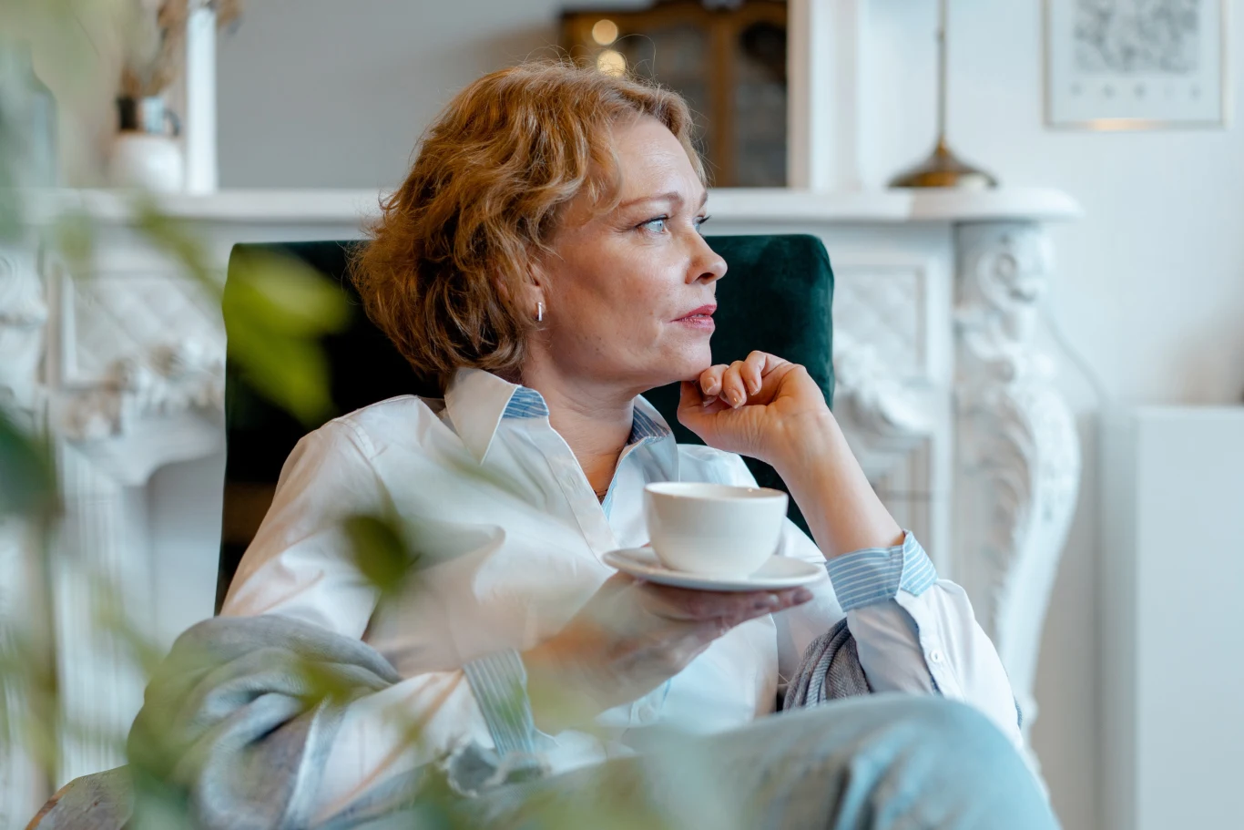 Kobieta o blond włosach w średnim wieku siedzi w fotelu z filiżanką herbaty lub kawy, patrzy zamyślona w bok, ubrana w jasną koszulę, w tle elegancki biały kominek.