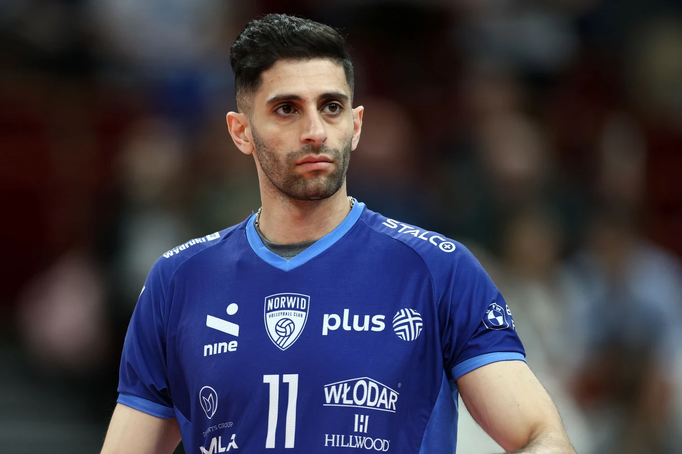 Milad Ebadipour, przyjmujący zespołu z Częstochowy Zawodnik siatkarski w niebieskiej koszulce z numerem 11, reprezentujący klub Norwid, z poważnym wyrazem twarzy, na tle rozmytej sali sportowej.