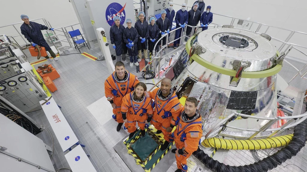 Cztery osoby w pomarańczowych skafandrach kosmicznych stoją w pobliżu kapsuły kosmicznej w pomieszczeniu technicznym NASA, w tle kilku pracowników w niebieskich uniformach i rękawicach.