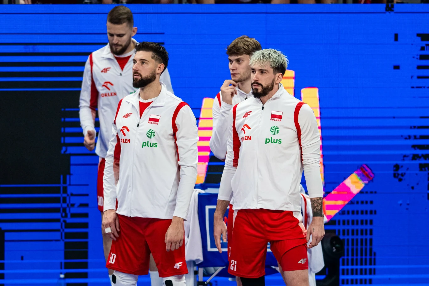 Bartosz Bednorz, Tomasz Fornal i Mateusz Poręba Polscy siatkarze w sportowych strojach reprezentacji narodowej na tle elektronicznej tablicy, skupieni przed rozpoczęciem meczu lub ważnym momentem spotkania.
