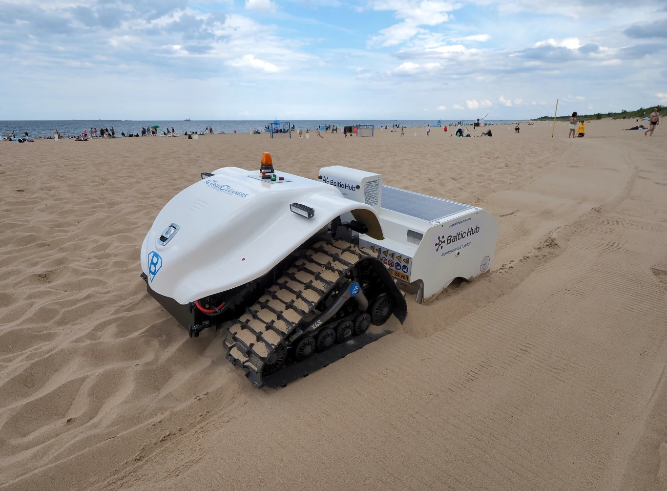 Cichy, szybki i zasilany słońcem. BeBot przeczesuje piasek w Gdańsku Robot gąsienicowy z białą obudową i logo Baltic Hub porusza się po piaszczystej plaży, w tle plażowicze wypoczywający nad morzem, przy częściowo zachmurzonym niebie.
