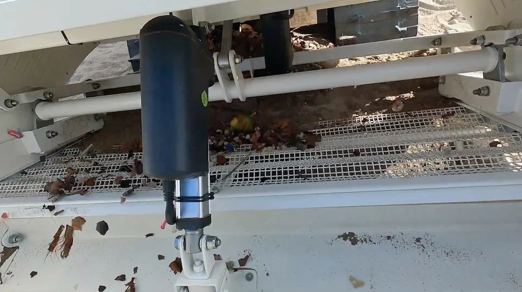 Część w pełni zmechanizowanej maszyny przemysłowej wyposażona w siatki filtrujące, metalowe pręty oraz czarny, cylindryczny element. Na siatkach widoczne są liście i drobne odpady.