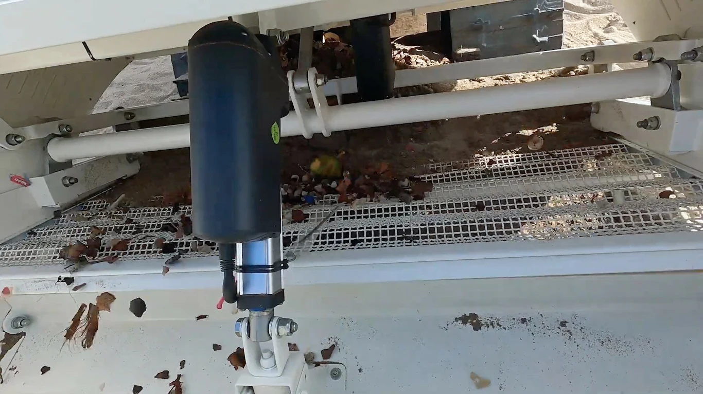 Śmieci trafiają do specjalnego pojemnika wewnątrz maszyny Część w pełni zmechanizowanej maszyny przemysłowej wyposażona w siatki filtrujące, metalowe pręty oraz czarny, cylindryczny element. Na siatkach widoczne są liście i drobne odpady.