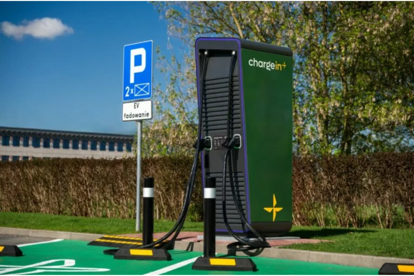 Stacja ładowania samochodów elektrycznych z widocznym znakiem informującym o miejscu parkingowym przeznaczonym do ładowania pojazdów. W tle krzewy i drzewa, a całość znajduje się na świeżym powietrzu.