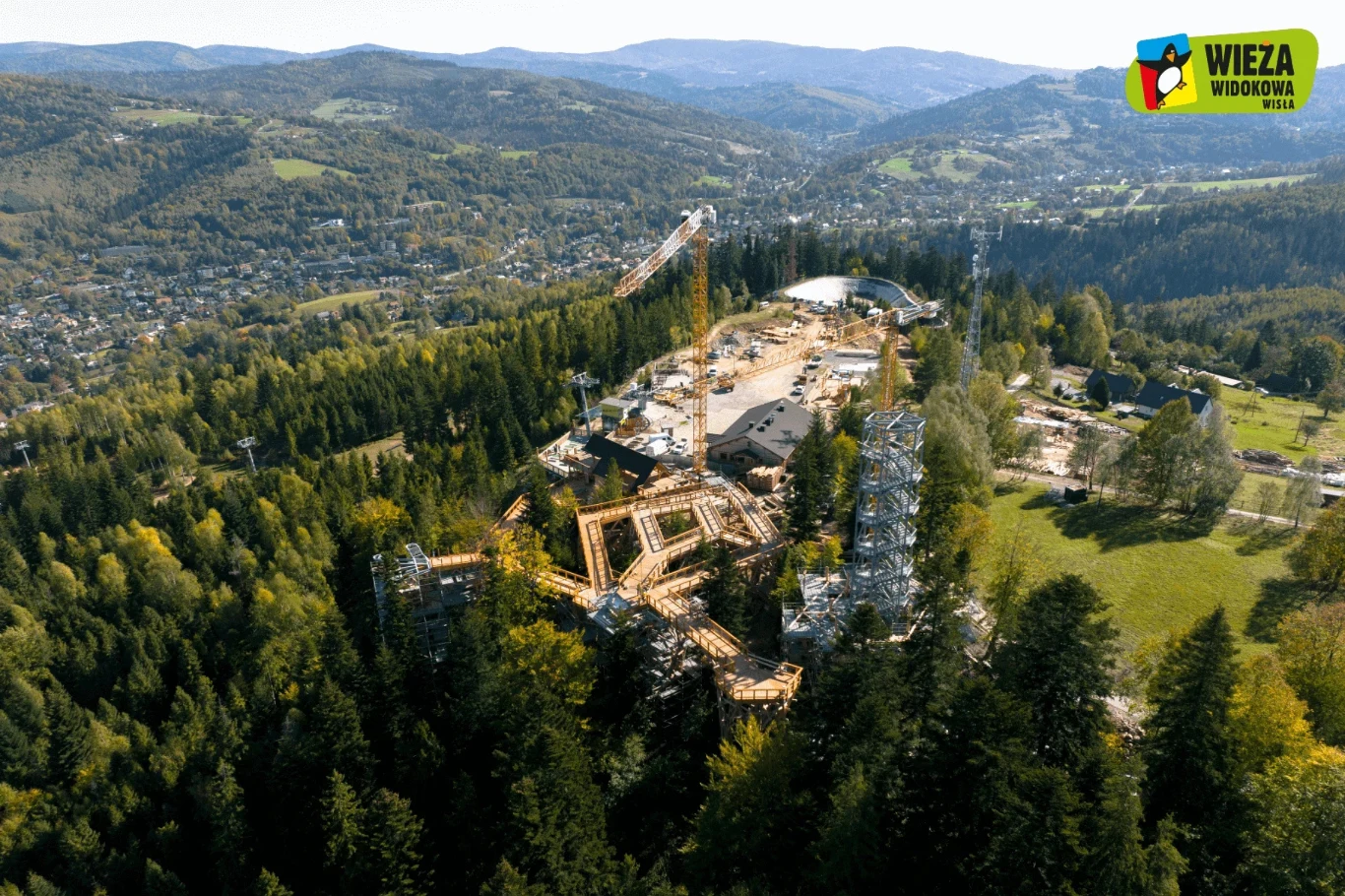 Konstrukcja wieży widokowej w otoczeniu górskich lasów, z lotu ptaka widoczne są rusztowania, maszyny budowlane oraz rozciągający się krajobraz Beskidów i miasteczek w dolinie.