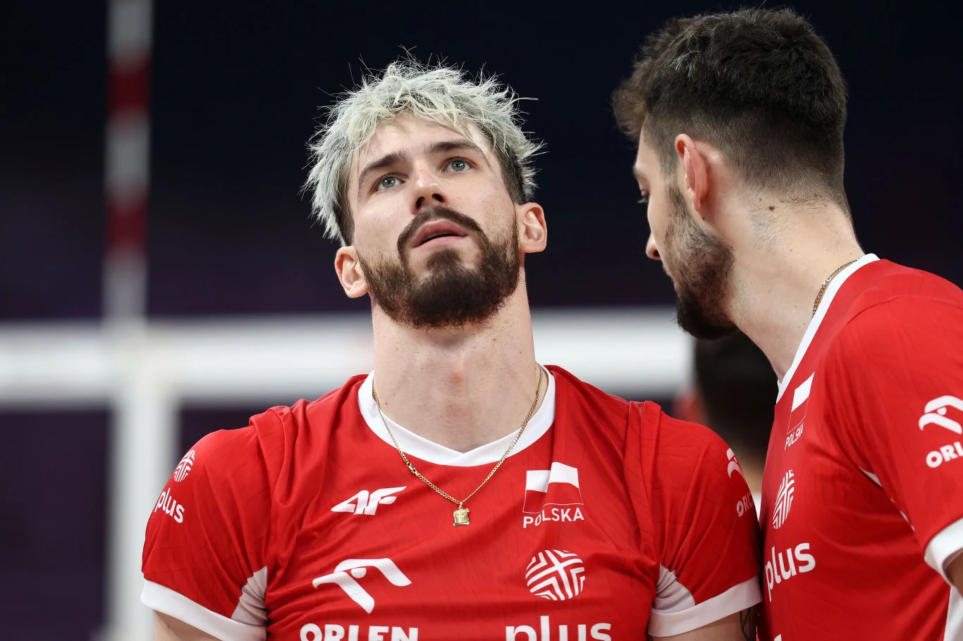 Dwaj siatkarze w czerwonych koszulkach reprezentacji Polski podczas meczu, jeden z nich z brodą i blond włosami patrzy do góry, a drugi zwraca się do niego, jakby przekazywał wskazówki lub rozmawiał.