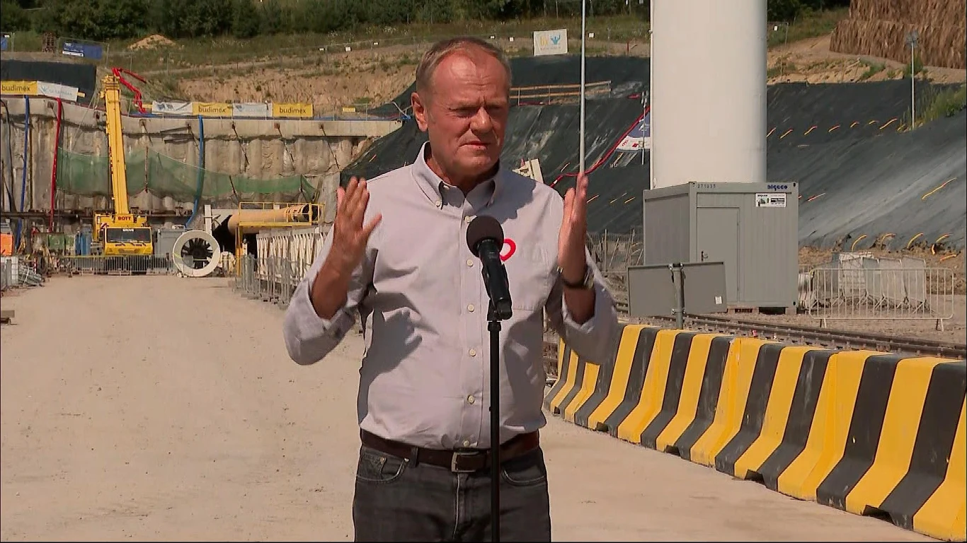 Donald Tusk odpowiedział na słowa szefa kancelarii prezydenta Mężczyzna w jasnej koszuli przemawia do mikrofonu na tle placu budowy, otoczony barierkami ochronnymi i ciężkim sprzętem budowlanym.